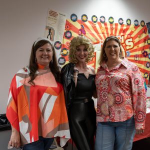 Angie Bersane, Dara Sanders-Aceves, Ruth D'Arezzo posing in front of their booth that is themed for the 1970's