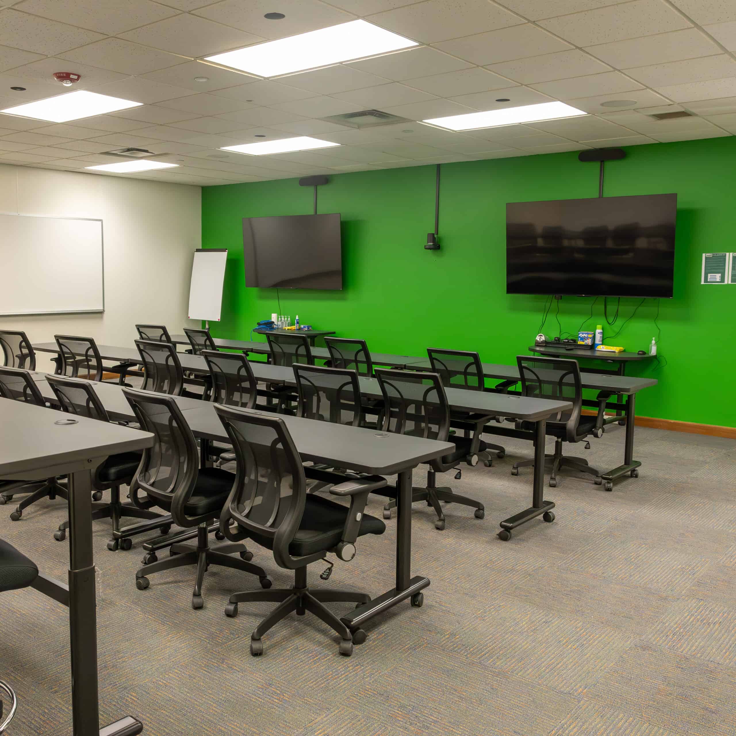 An interior photo of the ENMU-Roswell Learning Resource Center's "Flex Room", a multi-use space in room 121. A series of tables are arranged in rows with chairs positioned in front and facing toward the rear green wall where two large TVs are mounted.