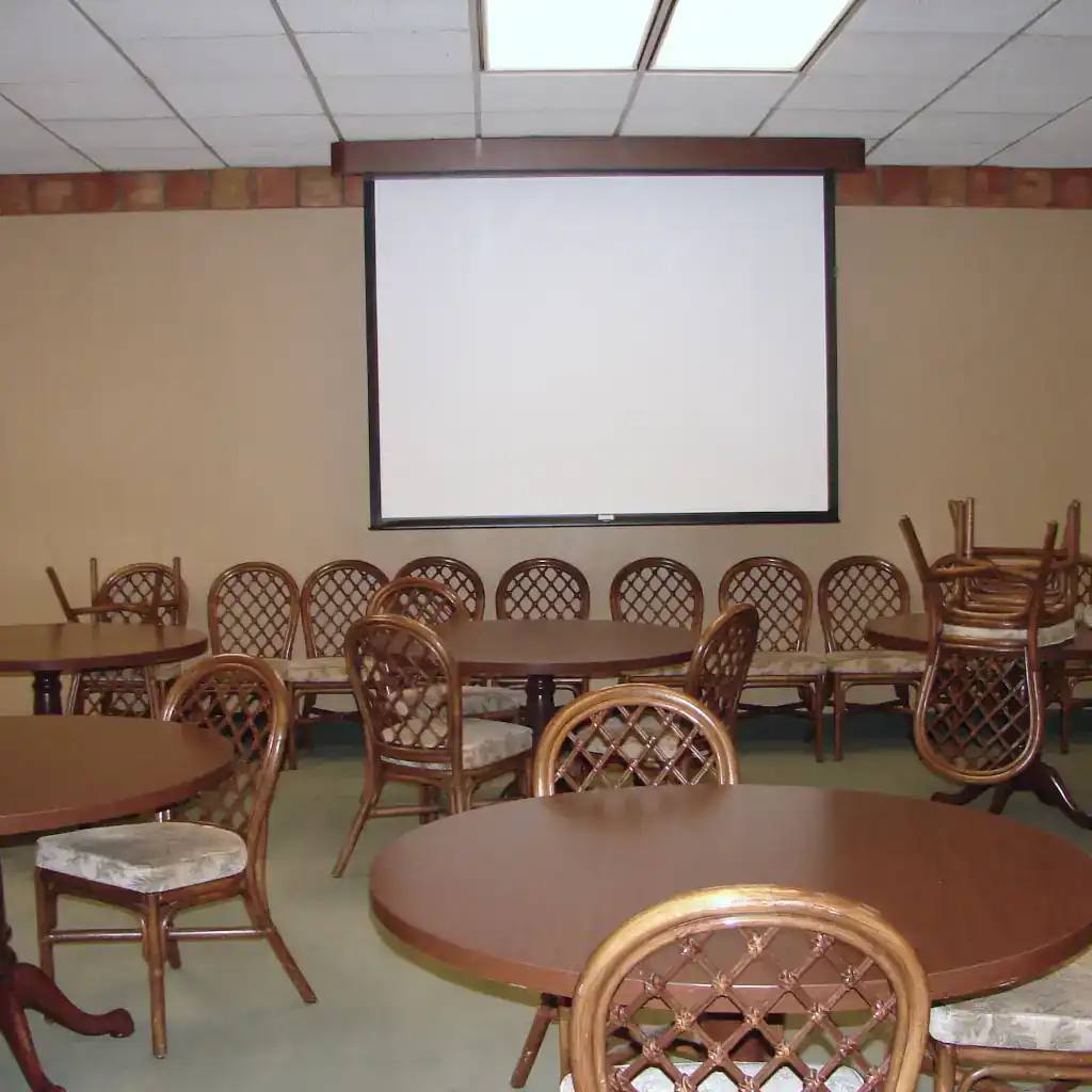 Photo of the CUB Alcove room. On the back wall hangs a projector screen. Many circular tables with chairs around them are in the foreground.