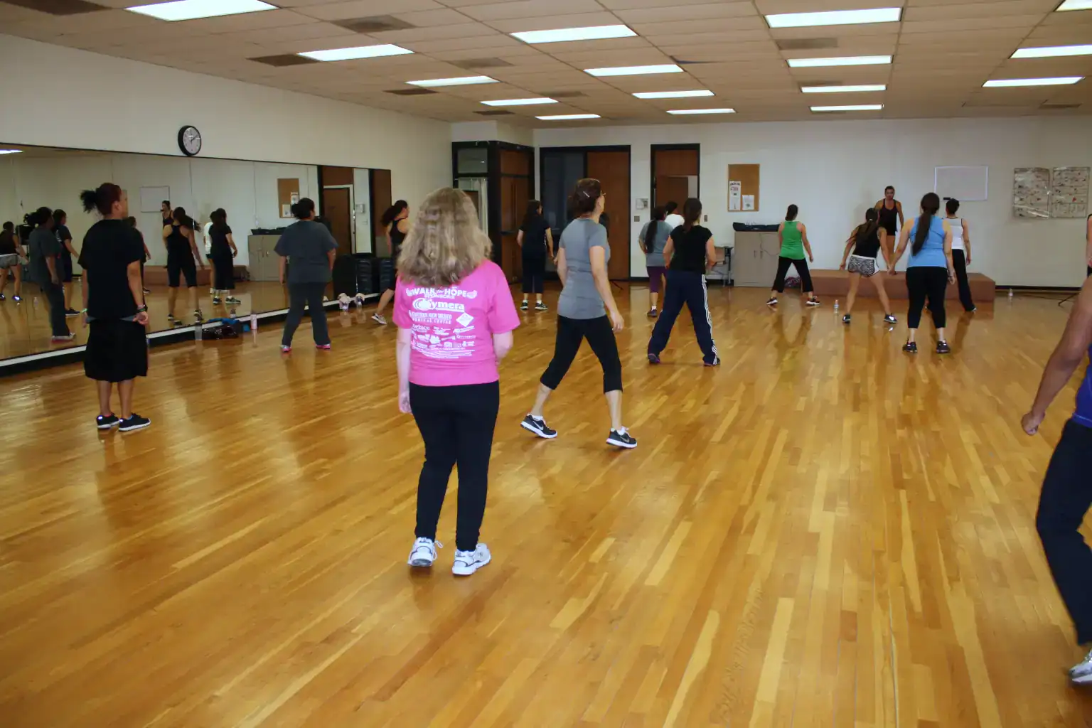 A photo of several people participating in a dance exercise on a wooden dance floor. A dance instructor leads the class in the background.