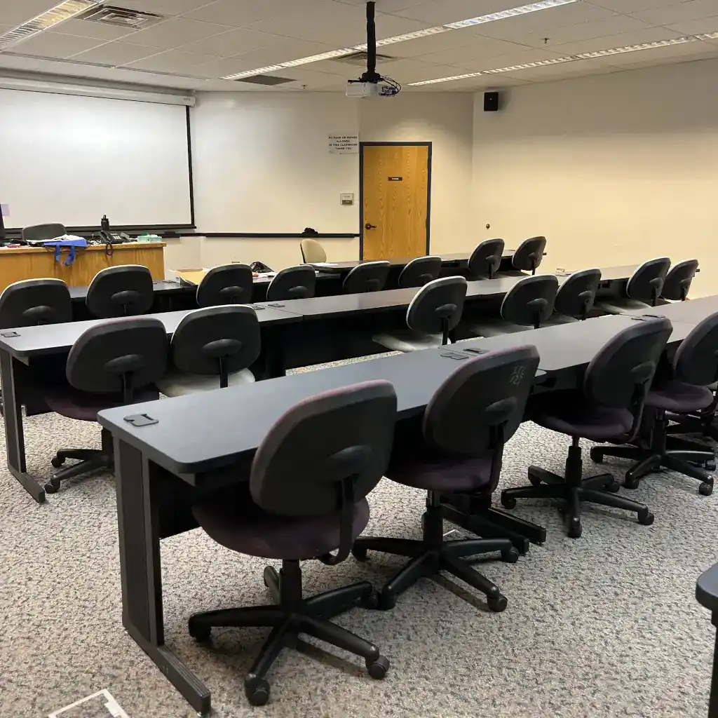Photo of a common non-computer lab classroom. Tables are set in rows with chairs docked behind them facing the front of the room where a projector screen is mounted.