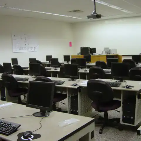 A photo of a classroom computer lab. There are four rows of tables each set with a row of chairs and a monitors.