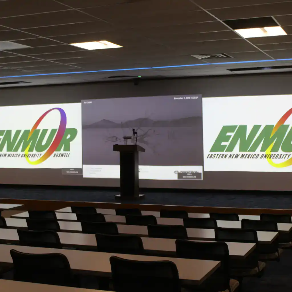 Several rows of tables and chairs face one large wall where three images are projected. In the center of the front stage is a podium.