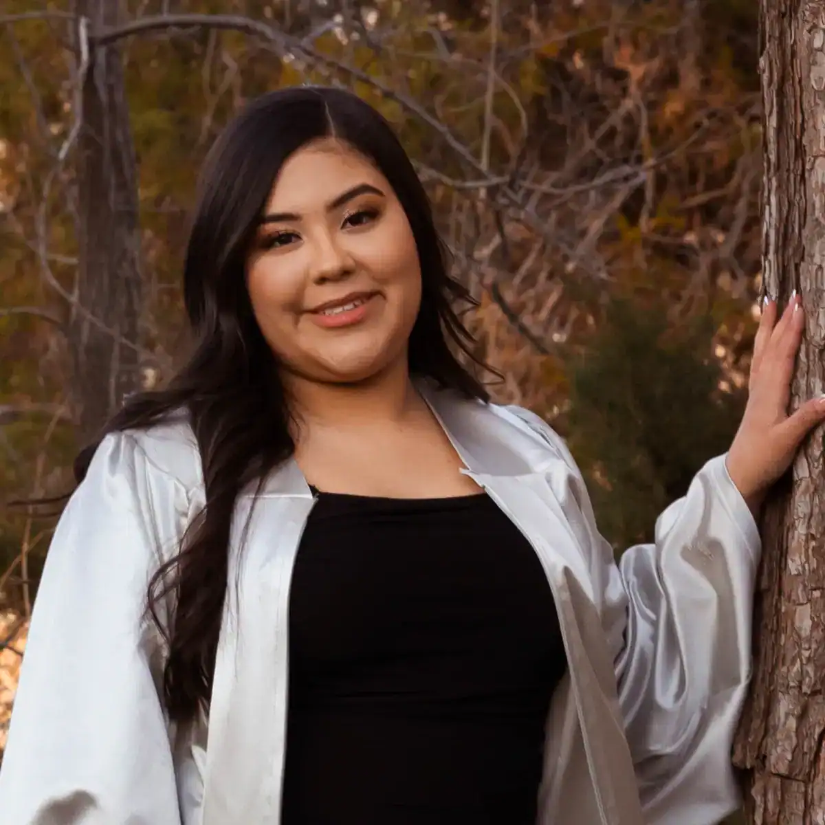 Photo of a smiling student in a graduation gown with one hand on the bark of a tree.