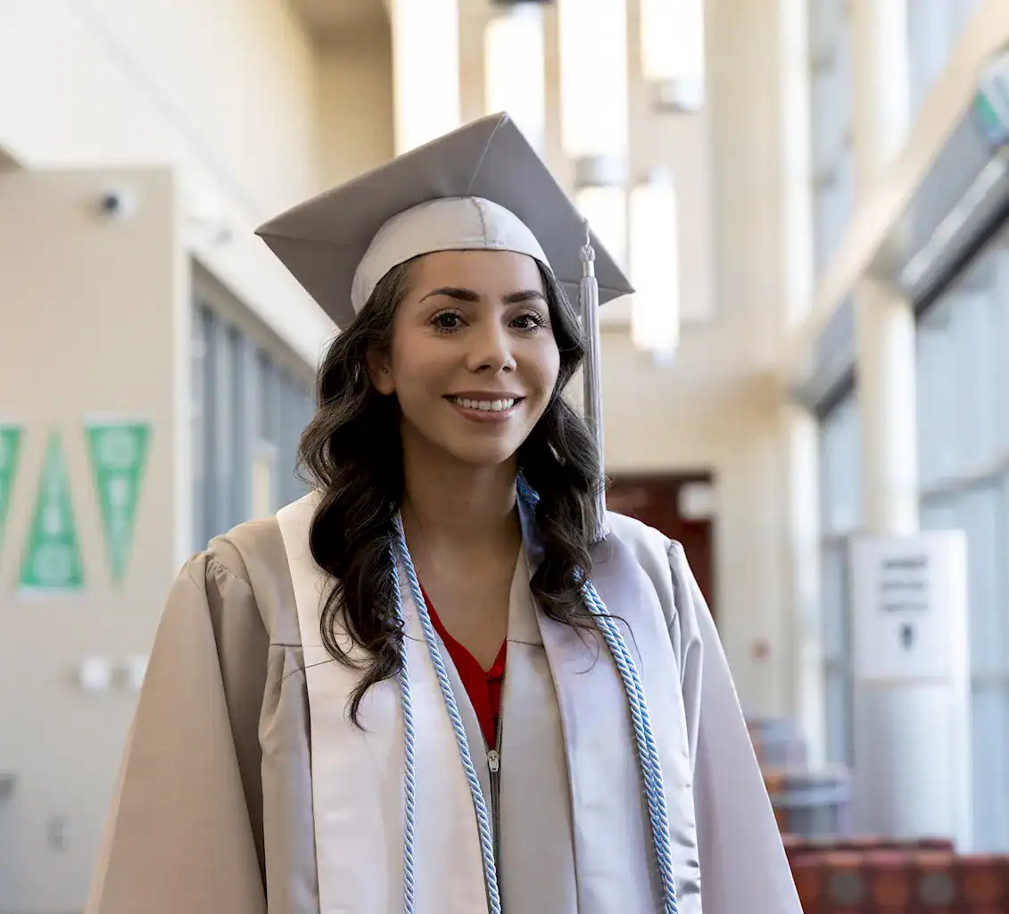 A photo of a smiling female graduate student in a white cap and gown standing inside the Student Services Center building.