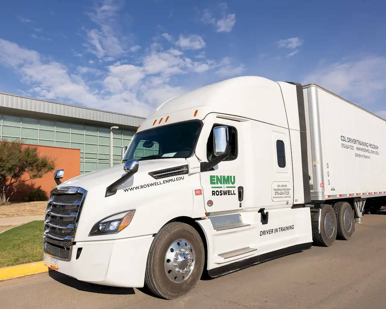 A colorful photo of a white semi truck parked in front of an orange building that has a glass facade. It is a bright sunny day, early to mid-morning. The sky is a rich blue with a few small high-altitude clouds floating in the background.<br />
The semi truck has the ENMU-Roswell logo printed on the driver's side door.