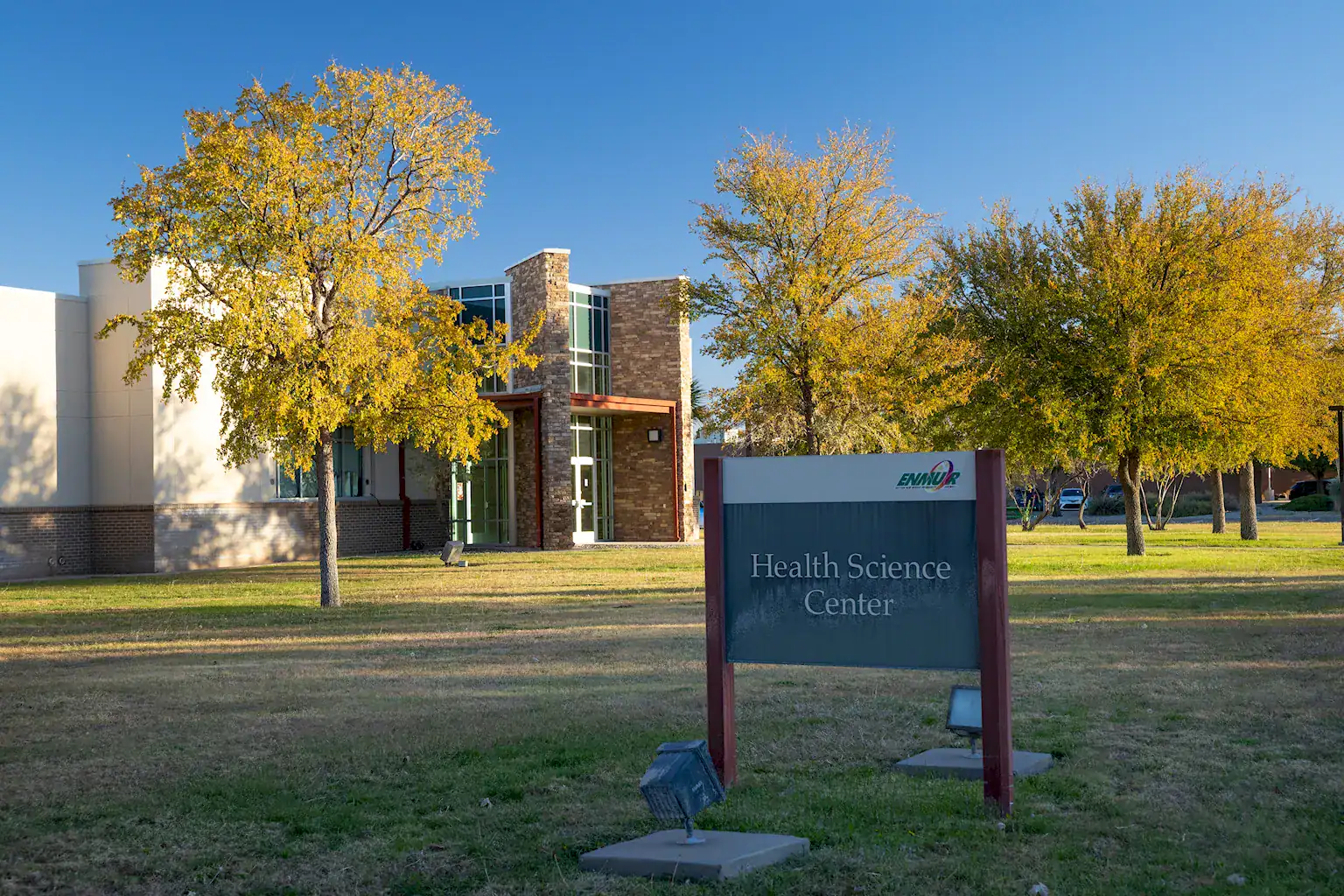 This image shows the exterior of the **Health Science Center** at the **ENMU-Roswell** campus on a clear, sunny day. ### **Key Features** * **The Building:** A modern, two-story structure featuring a mix of light-colored stucco, brick accents, and a prominent stone-pillared entrance with large glass windows. * **The Signage:** In the foreground, a dark teal monument sign with red posts reads **"Health Science Center"** in white text. The ENMUR logo is visible in the top right corner of the sign. * **Landscaping:** Several trees with vibrant yellow and green autumn foliage are scattered across a manicured green lawn. The sky in the background is a deep, clear blue. * **Lighting:** Bright, natural sunlight illuminates the scene from the left, casting soft shadows across the grass. Ground-mounted floodlights are positioned near the sign for nighttime illumination.