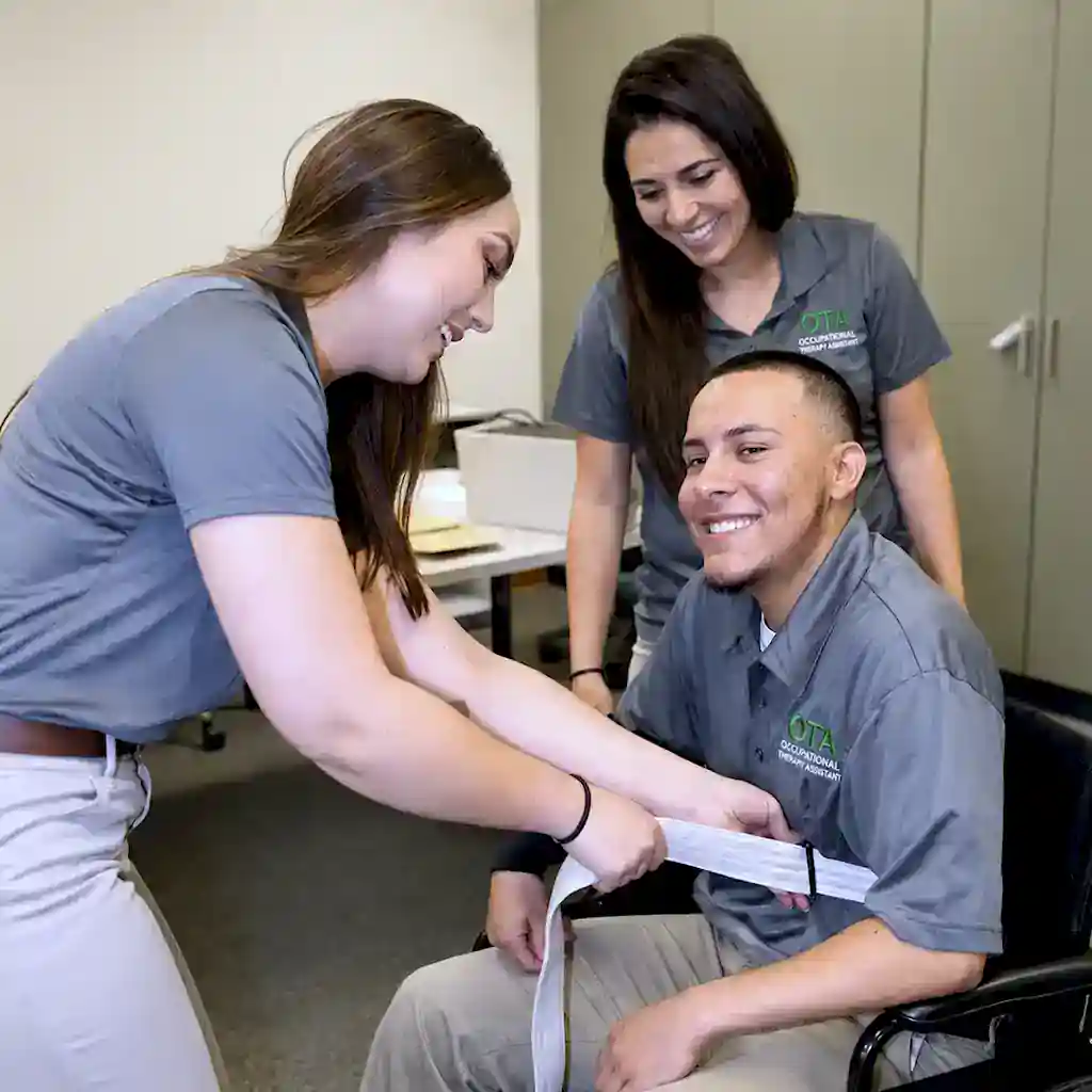 Occupational Therapy Assistant (OTA) students from ENMM-Roswell demonstrate securing a white strap around a man in a wheelchair during a clinical training session.